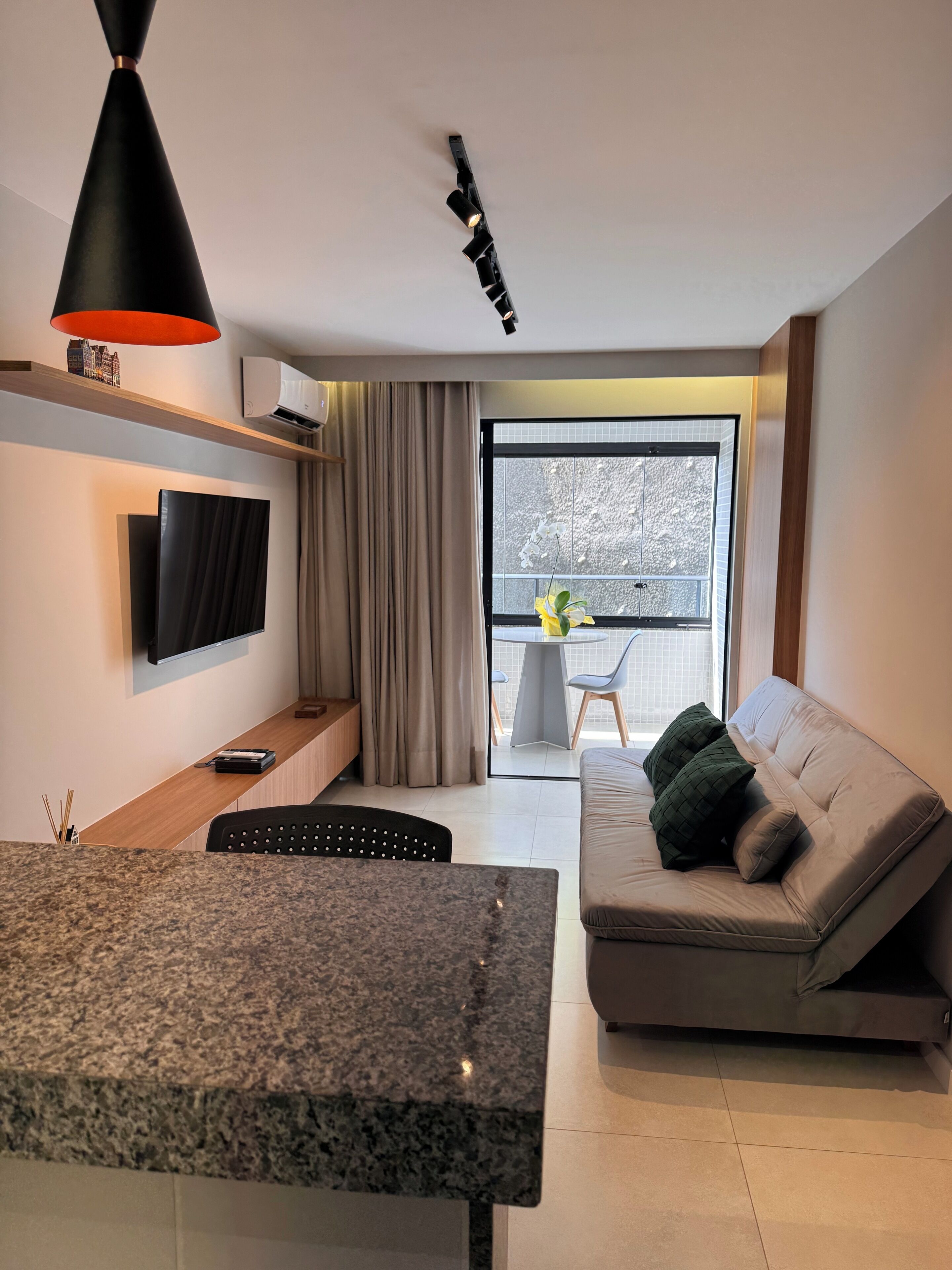 Un salon moderne doté d’un téléviseur à écran plat, d’un comptoir en granit, d’un canapé avec des coussins, d’un coin repas avec une table et d’une grande fenêtre offrant une vue sur les arbres.