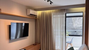 A modern living room with a flat-screen TV mounted on the wall, a wooden console table, a beige sofa with cushions, and a large window with sheer curtains. - Cozy NEW apartment in Ondina (Salvador)