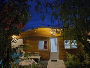 Una cabaña de madera con puerta y ventanas, rodeada de plantas y un banco.