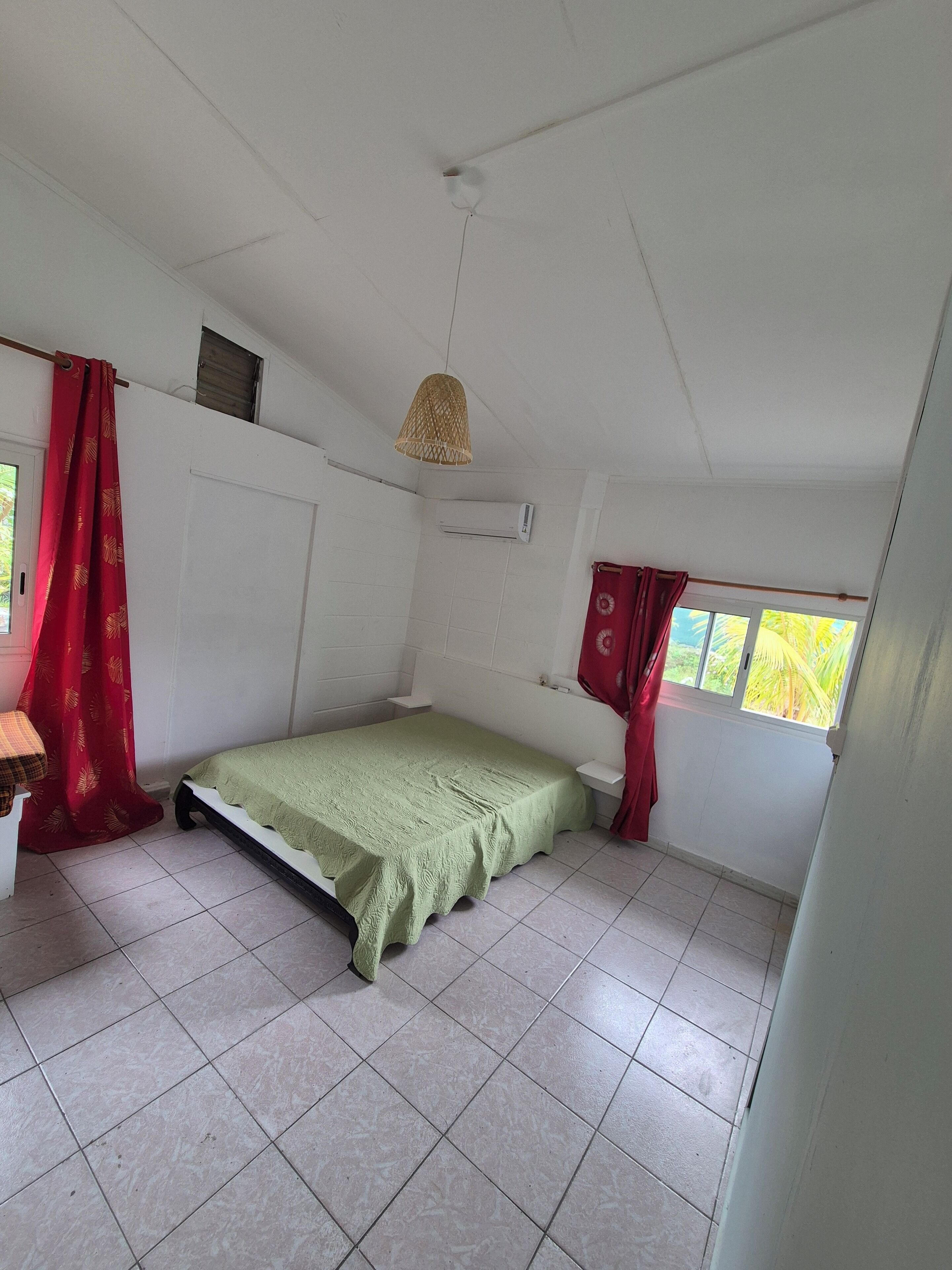 Quarto pequeno com cama de solteiro, coberta verde, paredes brancas e janela com cortinas vermelhas.