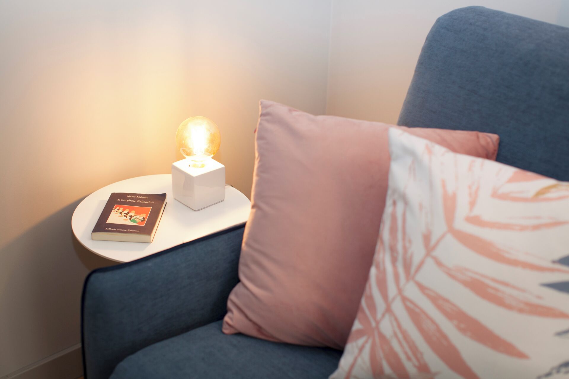Ein gemütlicher Winkel mit einem blauen Sofa, einem kleinen weißen Beistelltisch, einem Buch und einer beleuchteten Lampe.