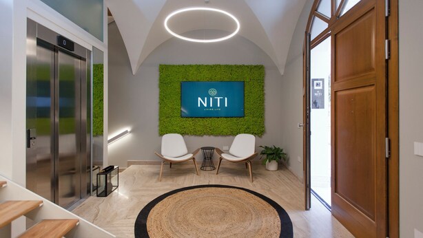 Ein modernes Interieur mit einer Rezeption, zwei weißen Stühlen, dem NITI-Logo an der Wand, einer Holztür und einer Treppe.