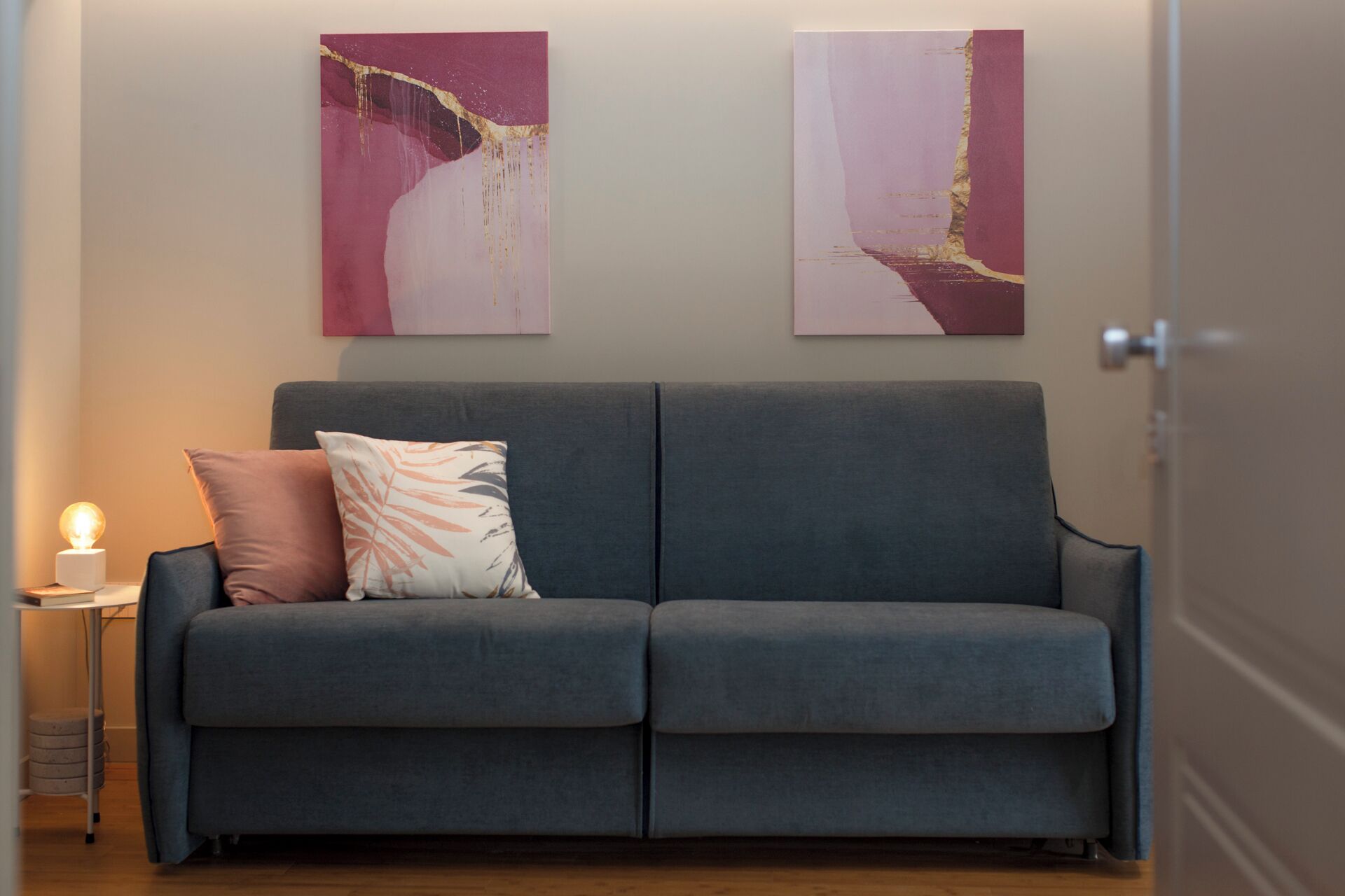 A modern living room with a grey sofa, a small side table with a lamp, and two abstract paintings on the wall.
