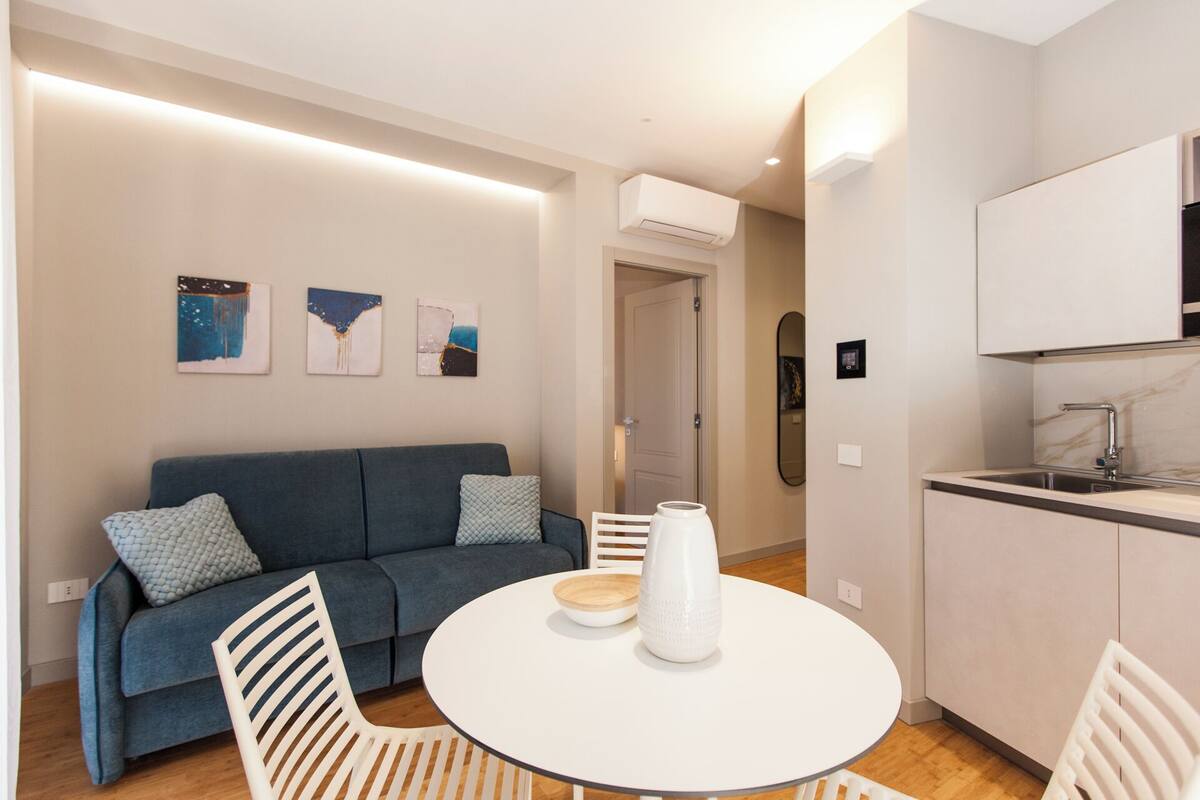 Una sala de estar moderna con un sofá azul, una mesa redonda blanca y una zona de cocina con un lavaplatos y armarios.