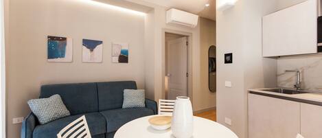 A modern living room with a blue sofa, a round white table, and a kitchen area with a sink and cabinets.