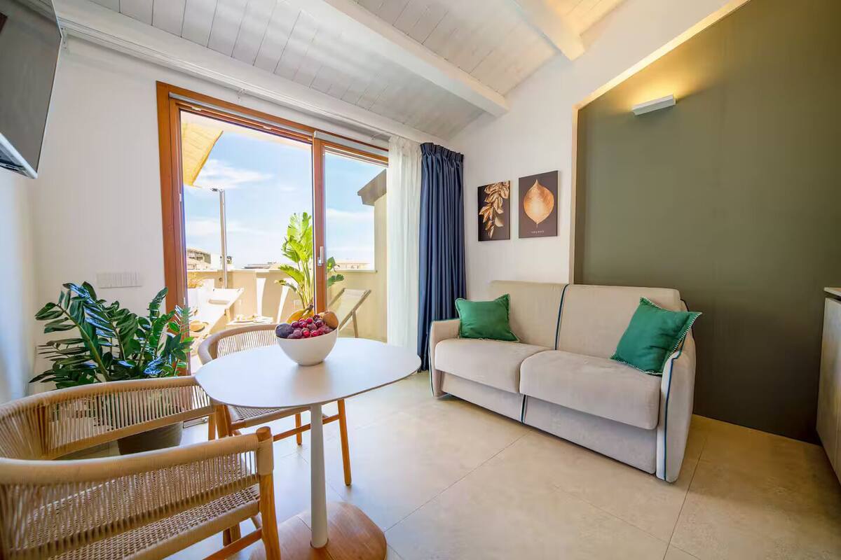 Una sala moderna con un sofá beige, una mesa redonda con un frutero, sillas de mimbre y vistas al exterior a través de ventanas amplias.
