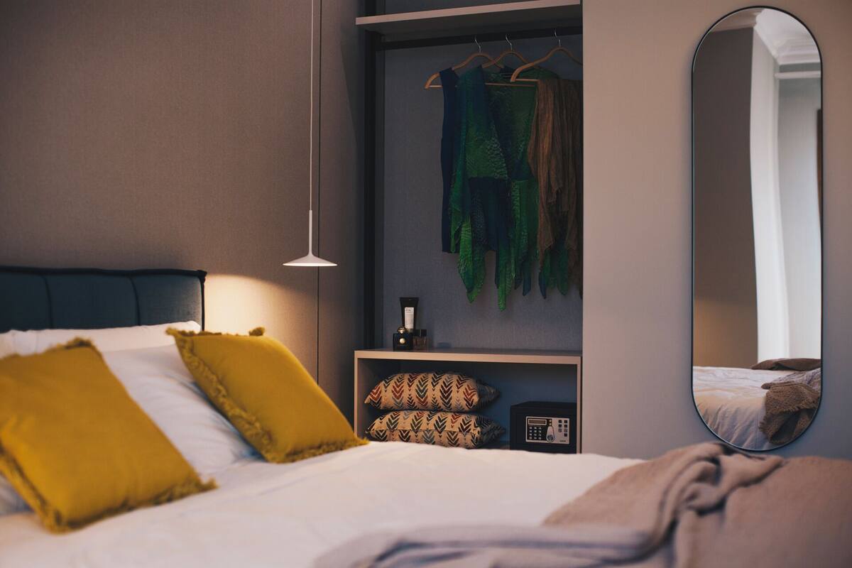 Un dormitorio con cama, almohadas, un perchero, un espejo y una pequeña estantería con artículos de aseo.