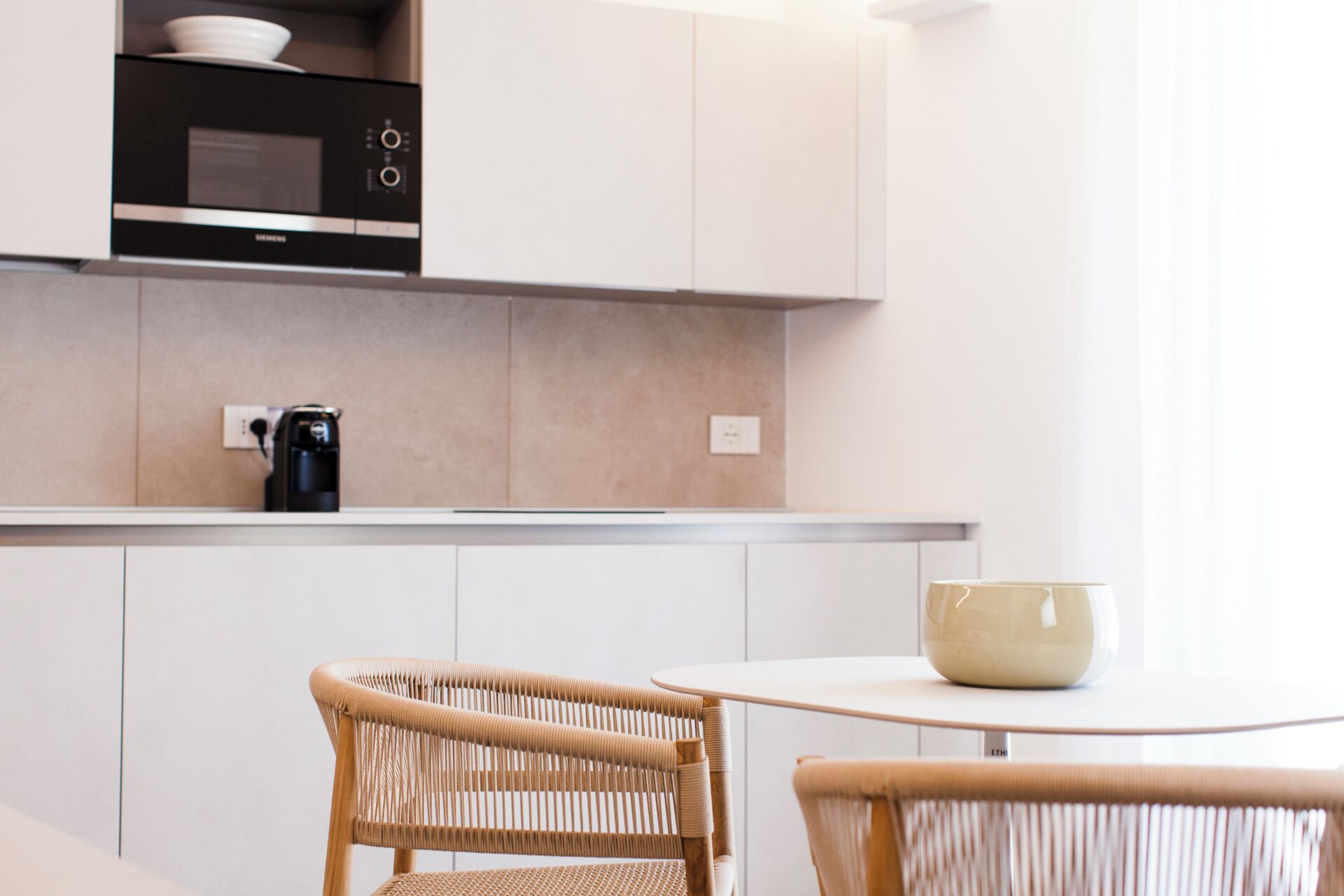 A modern kitchen with white cabinets, a black microwave, and a coffee maker on the counter. There is a round table with a beige bowl on it, and two wicker chairs.