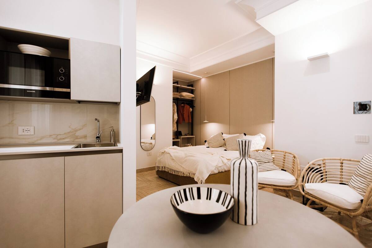 Un dormitorio moderno y bien iluminado con una cama, mesitas de noche y un espejo grande. Junto a él, una kitchenette con un microondas y un horno Siemens.