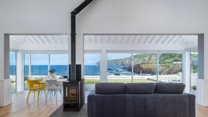 Ein geräumiges Wohnzimmer mit großem Fenster und Blick auf die Küste, einem Kamin und einem grauen Sofa.