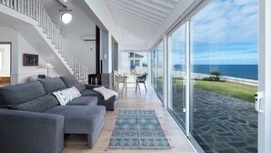 Ein modernes Wohnzimmer mit einem grauen Ecksofa, einem Essbereich mit Tisch und Stühlen sowie einer großen Glasdoor, die auf eine Terrasse mit Meerblick führt.