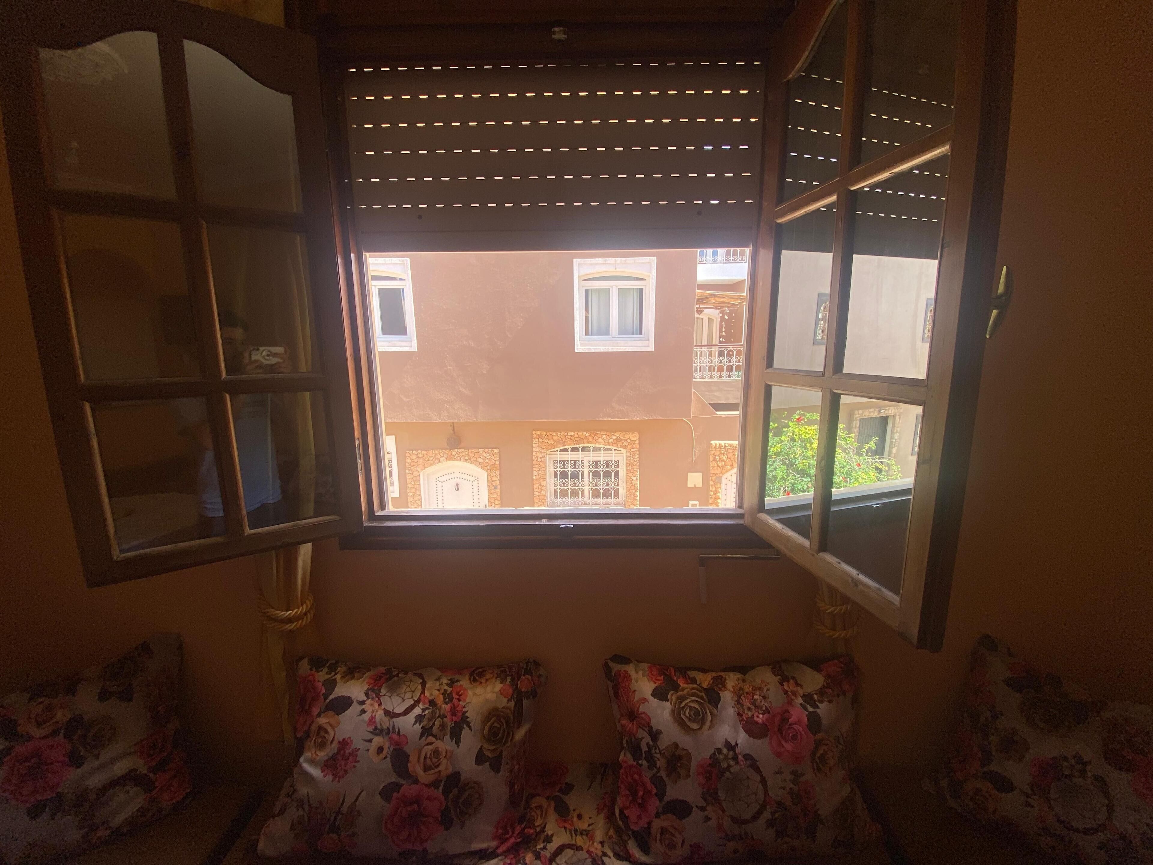 Une pièce dotée d’une grande fenêtre, d’un canapé agrémenté de coussins à motifs floraux et offrant une vue sur un immeuble avec des balcons.