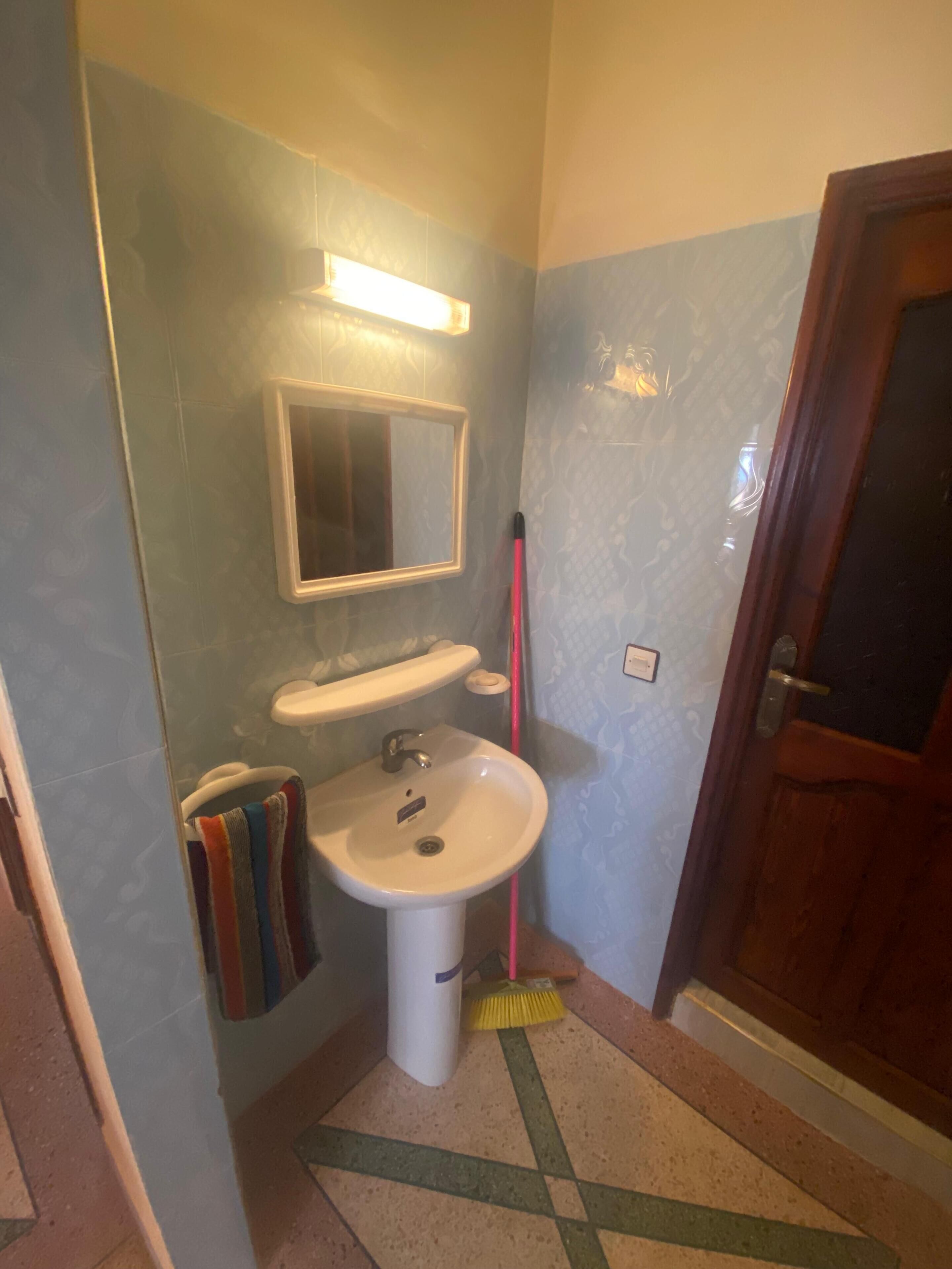 A bathroom with a sink, mirror, and a broom leaning against the wall.