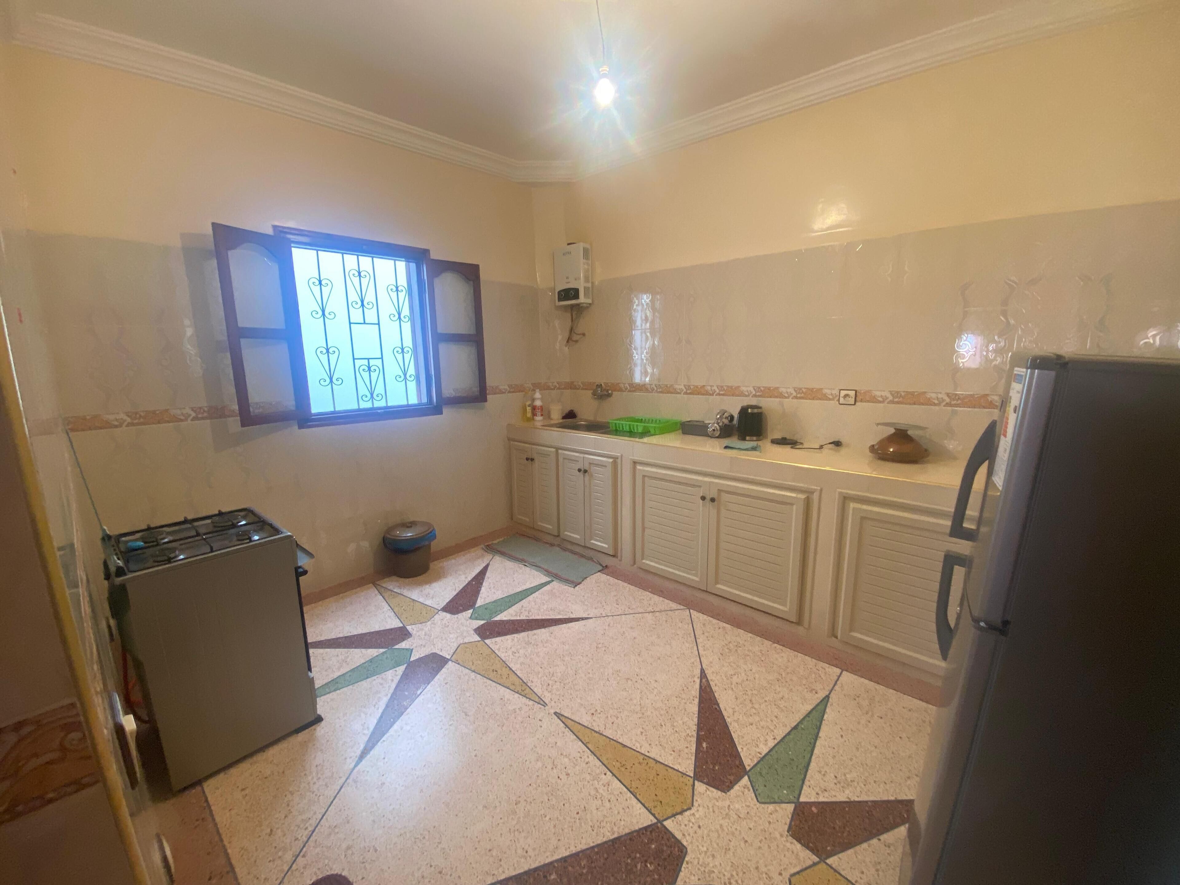 Una cocina con estufa a gas, un bote de basura, un refrigerador y una ventana con celosías decorativas.