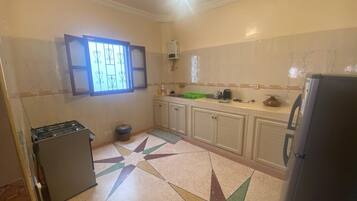 Una cocina con estufa a gas, un bote de basura, un refrigerador y una ventana con celosías decorativas.