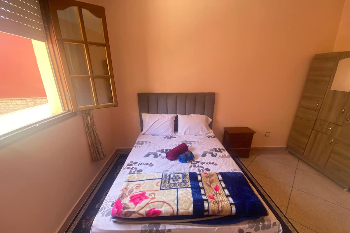 A neatly made bed with a floral-patterned bedspread and pillows, a small bedside table with a lamp, and a wardrobe in a room with a tiled floor and a window with blinds.