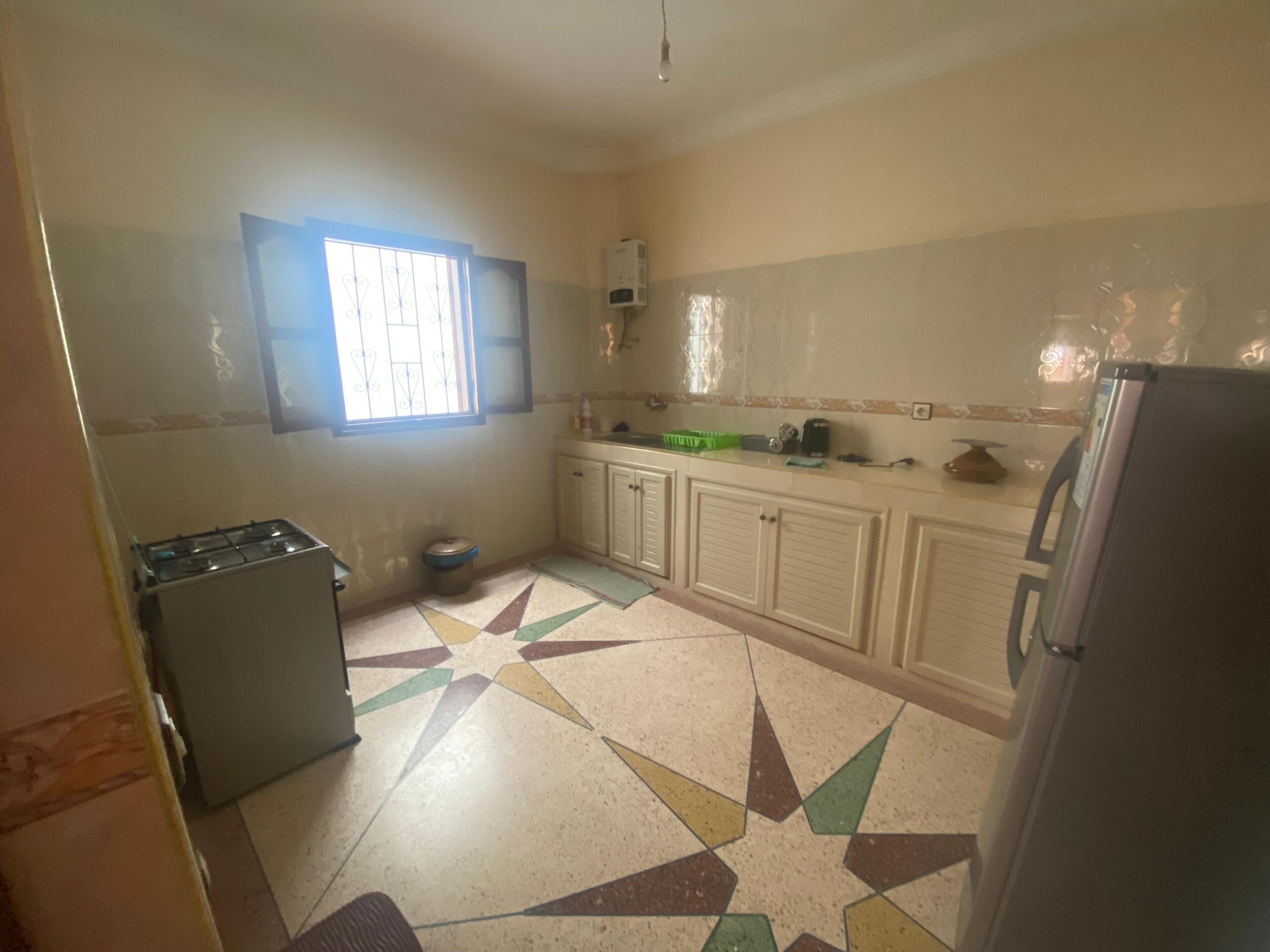 A kitchen with a gas stove, a refrigerator, cabinets, and a window with bars.