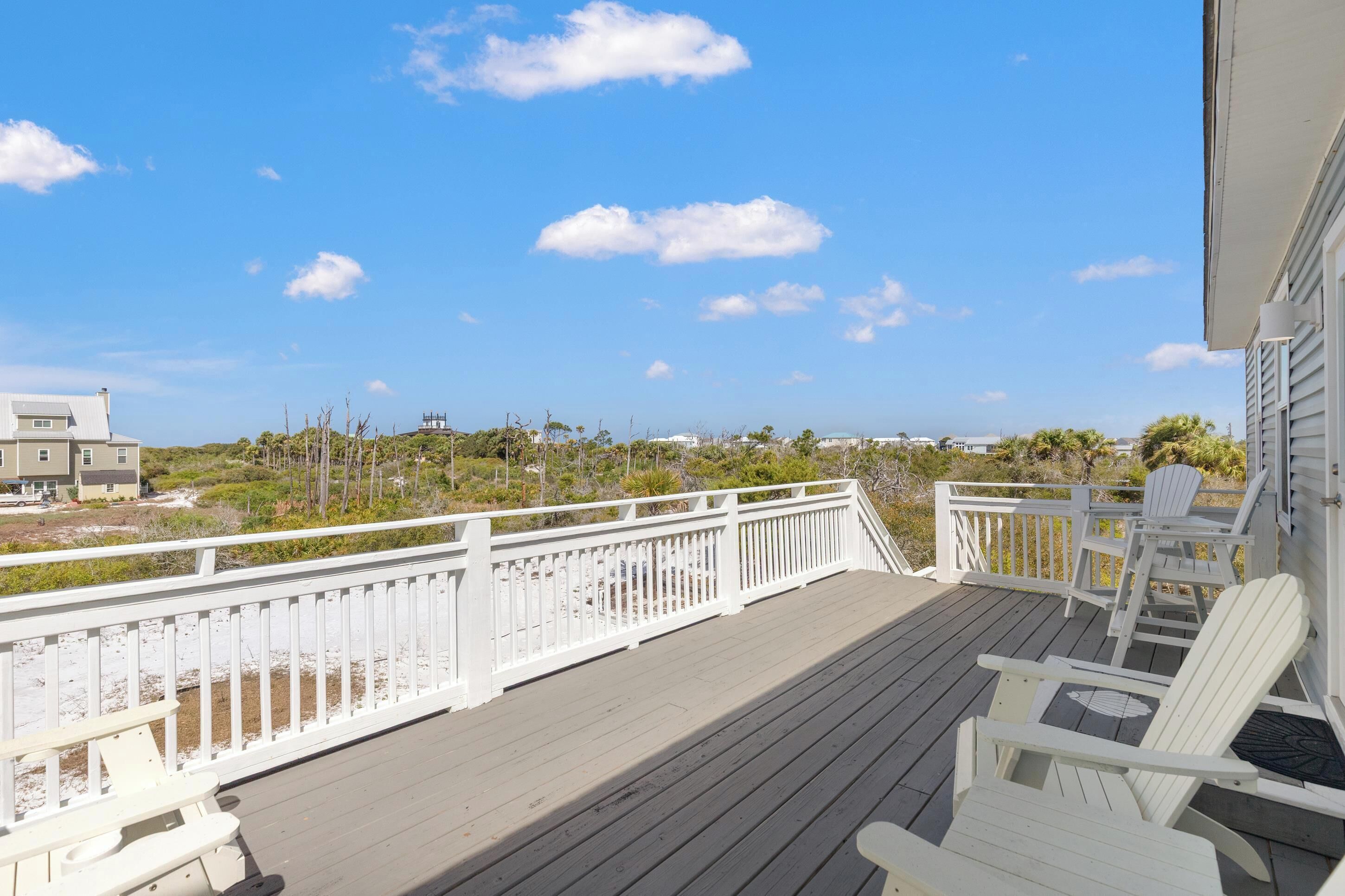Une terrasse en bois avec des balustrades blanches, des fauteuils Adirondack blancs et une vue sur la plage et l’océan.