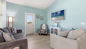 A living room with a grey sofa, a patterned armchair, a white side table, and a painting of a beach scene. - Beach House - Salty Dolphin (Port St. Joe)
