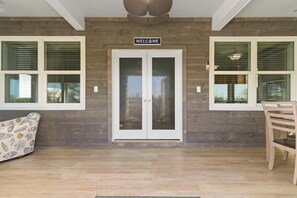 A wooden exterior wall with a 'WELCOME' sign above a double door, two windows with blinds, and a wooden chair to the right. - Beach House - Salty Dolphin (Port St. Joe)
