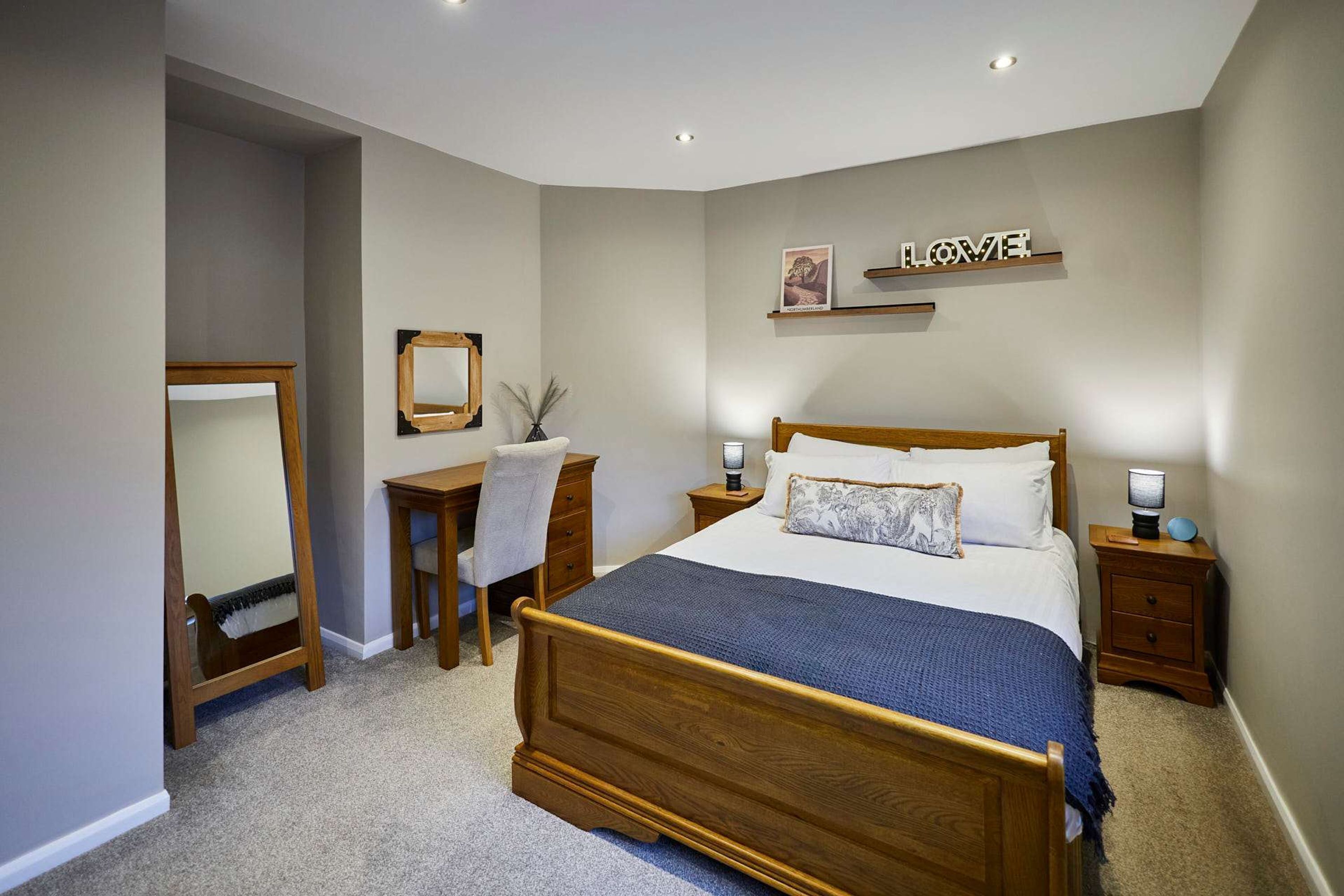 A bedroom with a wooden bed, a desk, a chair, a mirror, and a wall-mounted shelf with a picture.