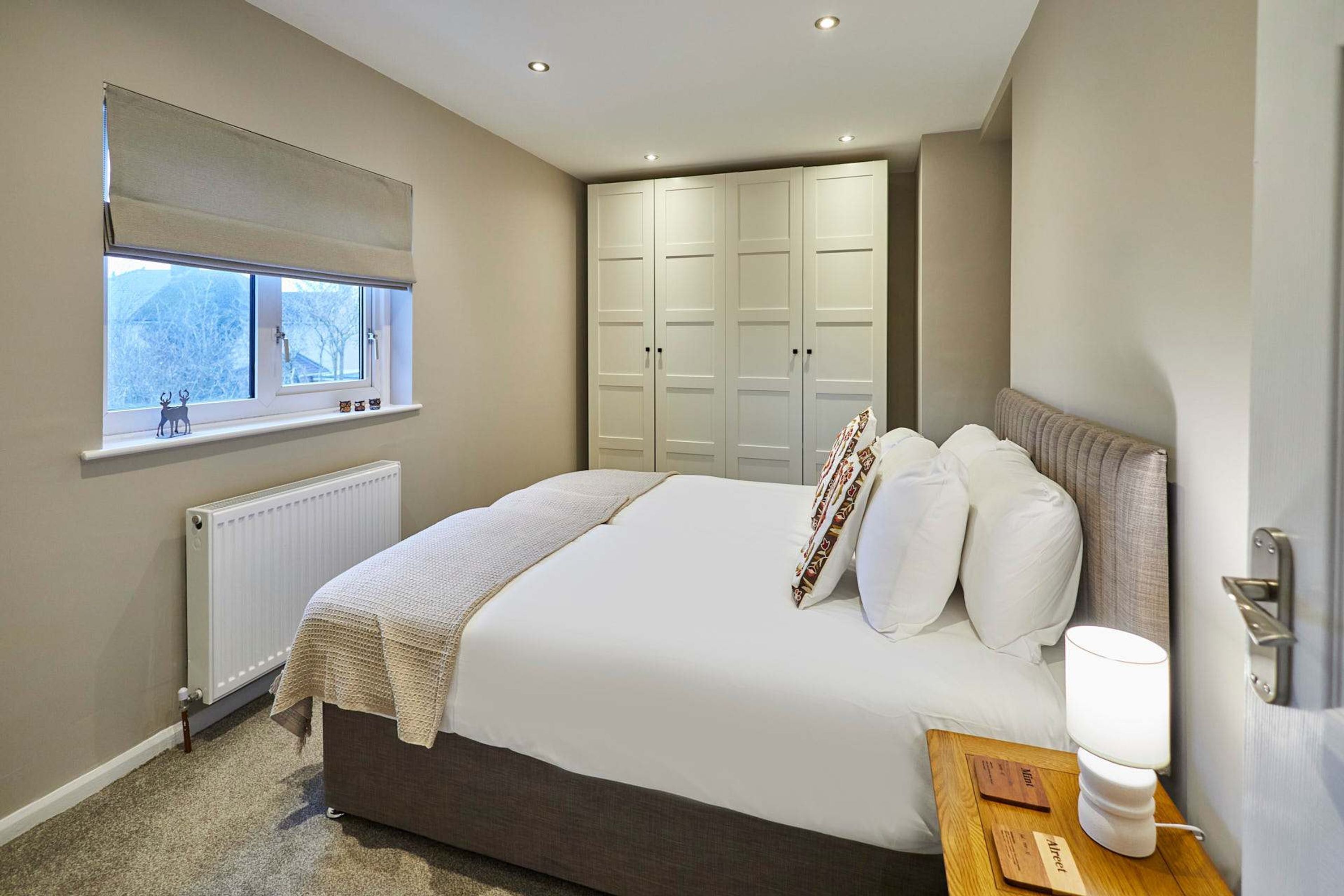 A bedroom with a large bed, a bedside table with a lamp, a window with a blind, and a wardrobe.