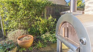 Ein metallener Grill für den Außenbereich mit Schornstein, ein Holzpflanzgefäß mit Grünpflanzen und ein Backsteinhaus im Hintergrund.
