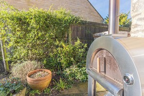 Ein metallener Grill für den Außenbereich mit Schornstein, ein Holzpflanzgefäß mit Grünpflanzen und ein Backsteinhaus im Hintergrund.