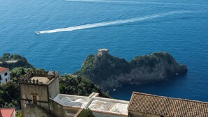 Vista costera con un edificio histórico, una isla rocosa y dos barcos dejando estelas en el agua.