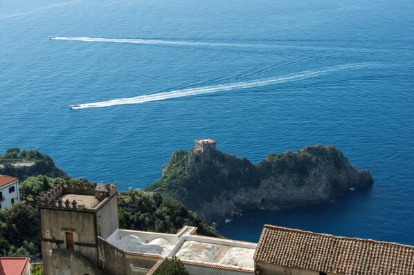 Vista costiera con un edificio storico, un'isola rocciosa e due imbarcazioni che creano scie nell'acqua.