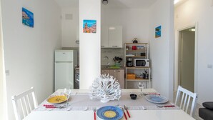Un comedor con una mesa dispuesta para cuatro personas, un refrigerador blanco, un microondas y un estante con utensilios de cocina.