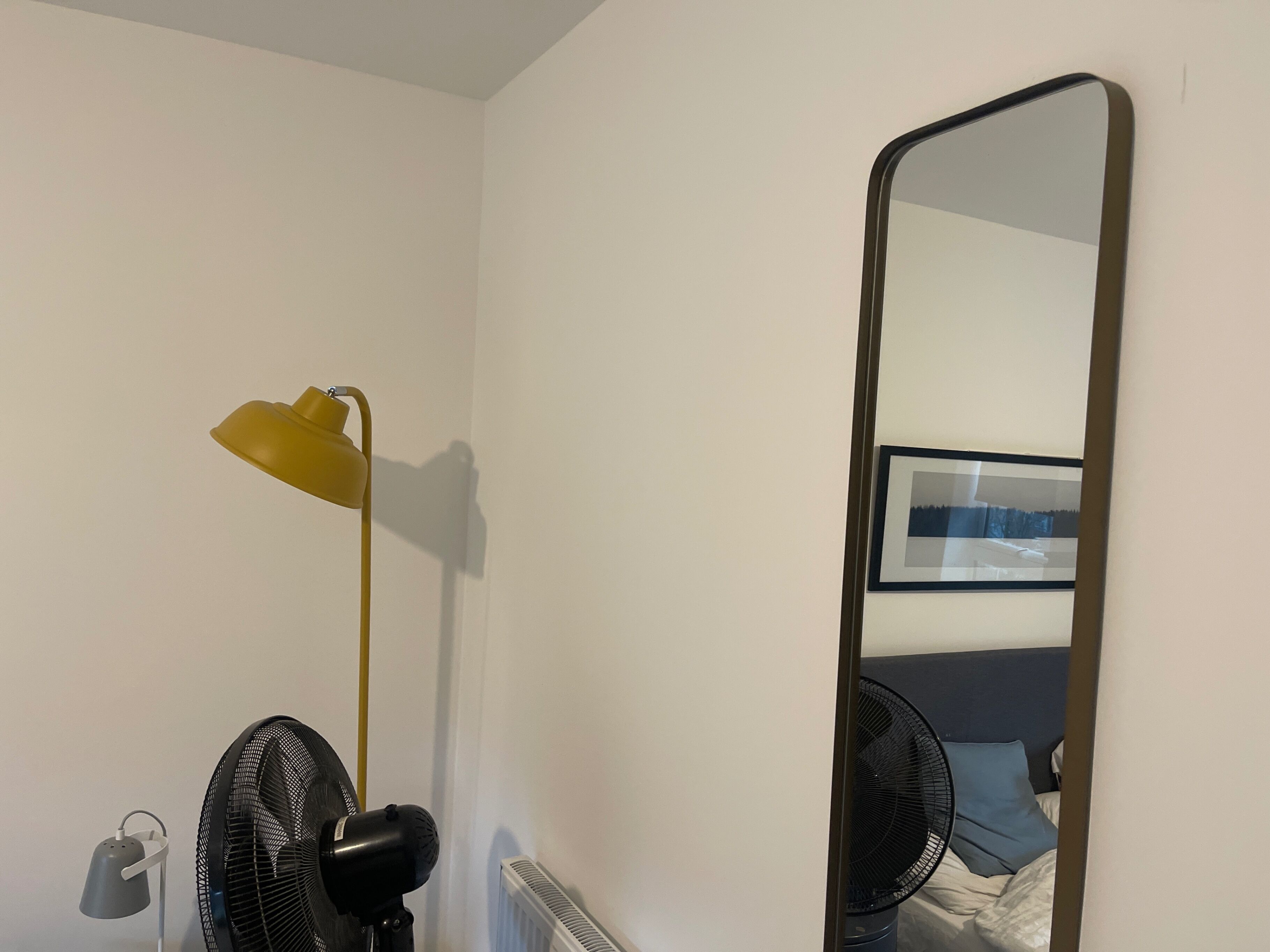 A room with a bed, a standing fan, a desk lamp, and a large mirror.
