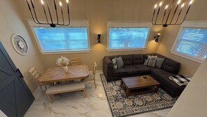 Ein Wohnzimmer mit Esstisch, einer Couch und einem Couchtisch. Zwei Fenster mit Jalousien, eine Chandelüster und ein dekoratives Wandbild sind ebenfalls zu sehen.