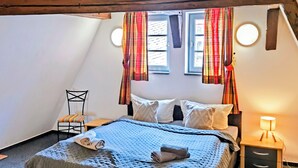 Un dormitorio acogedor con techo inclinado, una cama con colcha azul, dos almohadas, una silla y una mesita de noche con lámpara.