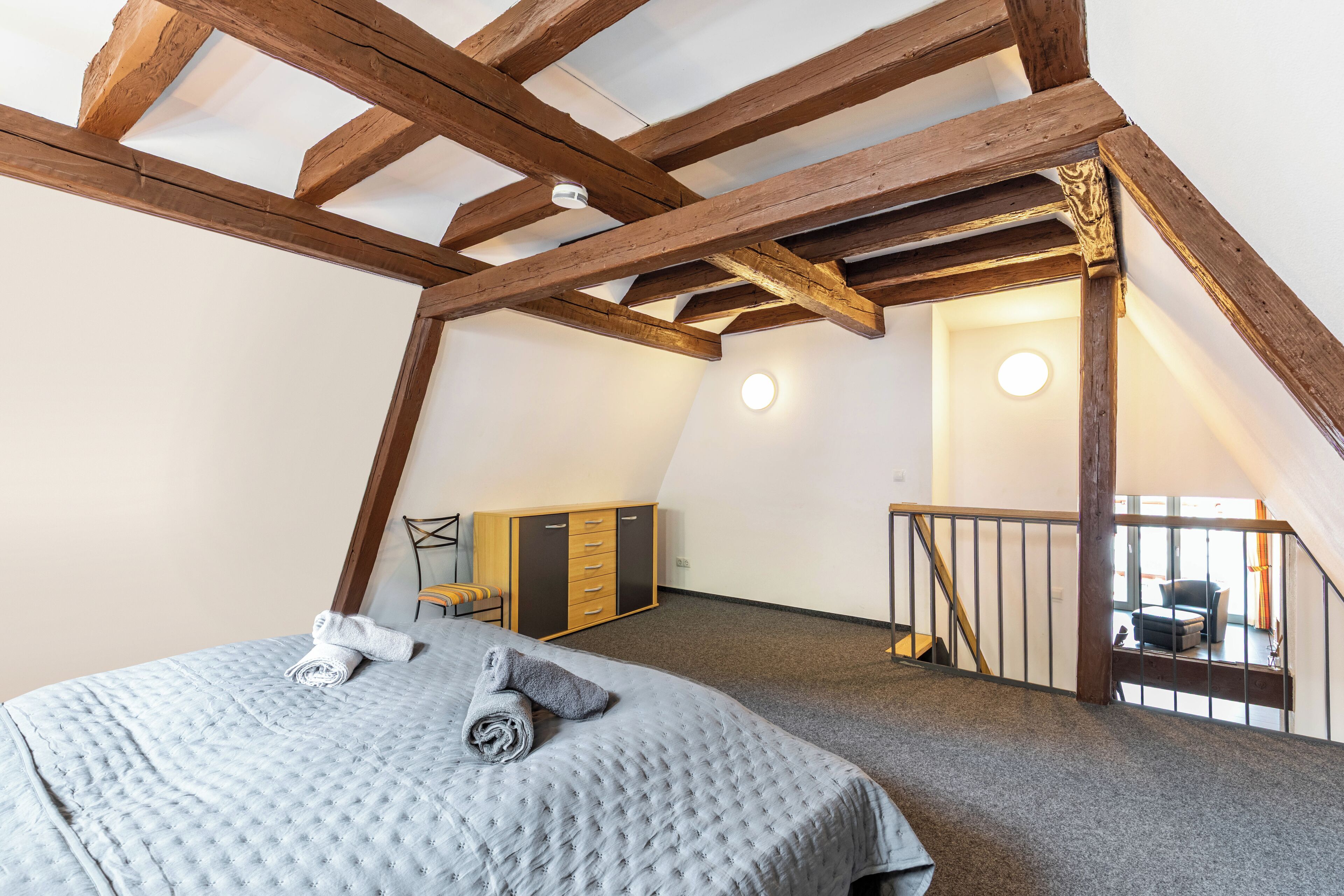 A bedroom with a high ceiling, exposed wooden beams, a bed with a quilt, a wardrobe, and a staircase leading to an upper level.