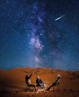 Escena desértica con tres personas sentadas a una mesa bajo un cielo estrellado.