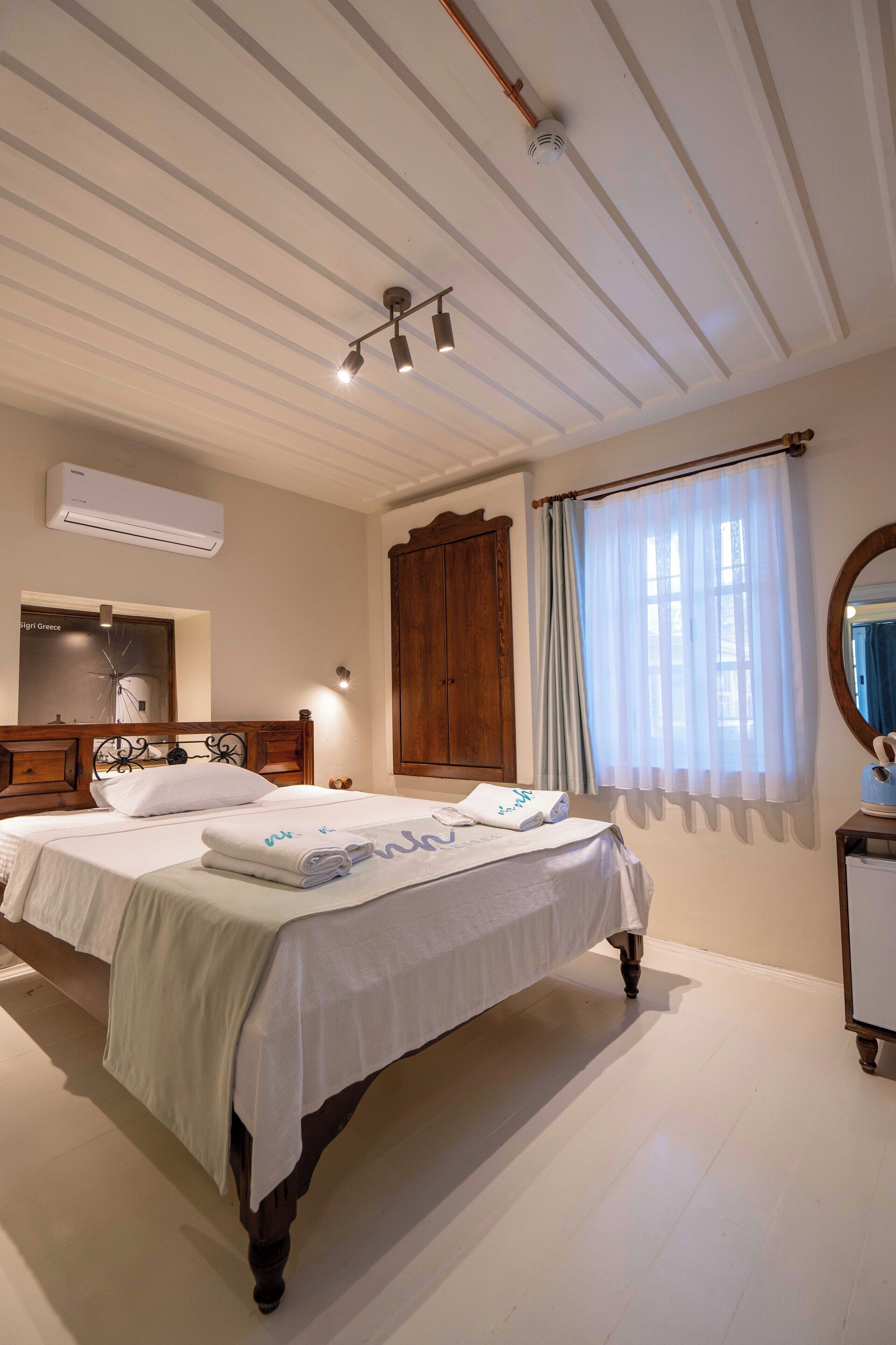 A well-lit bedroom with a large bed, a wooden wardrobe, an air conditioning unit, and a window with curtains.