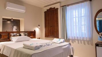 A well-lit bedroom with a large bed, a wooden wardrobe, an air conditioning unit, and a window with curtains.