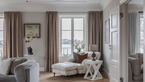 A living room with a sofa, ottoman, and a side table with a lamp. There is a window with curtains and a painting on the wall. - Villa Strandliv invites you to experience an extraordinary stay in a one-of-a-kind residence. (Gustavsberg)