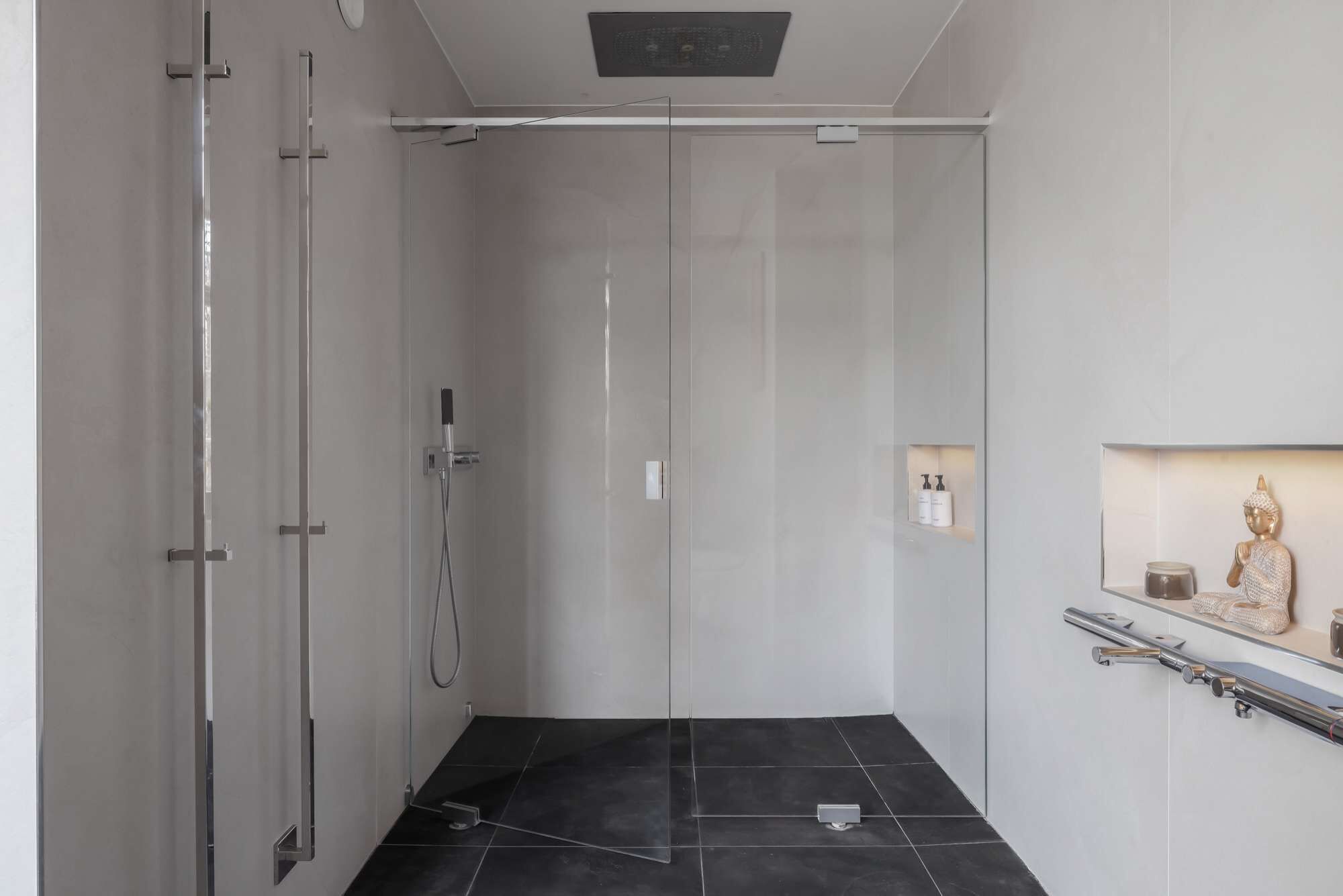 A modern bathroom with a glass shower enclosure, a black tiled floor, and a small shelf with toiletries.