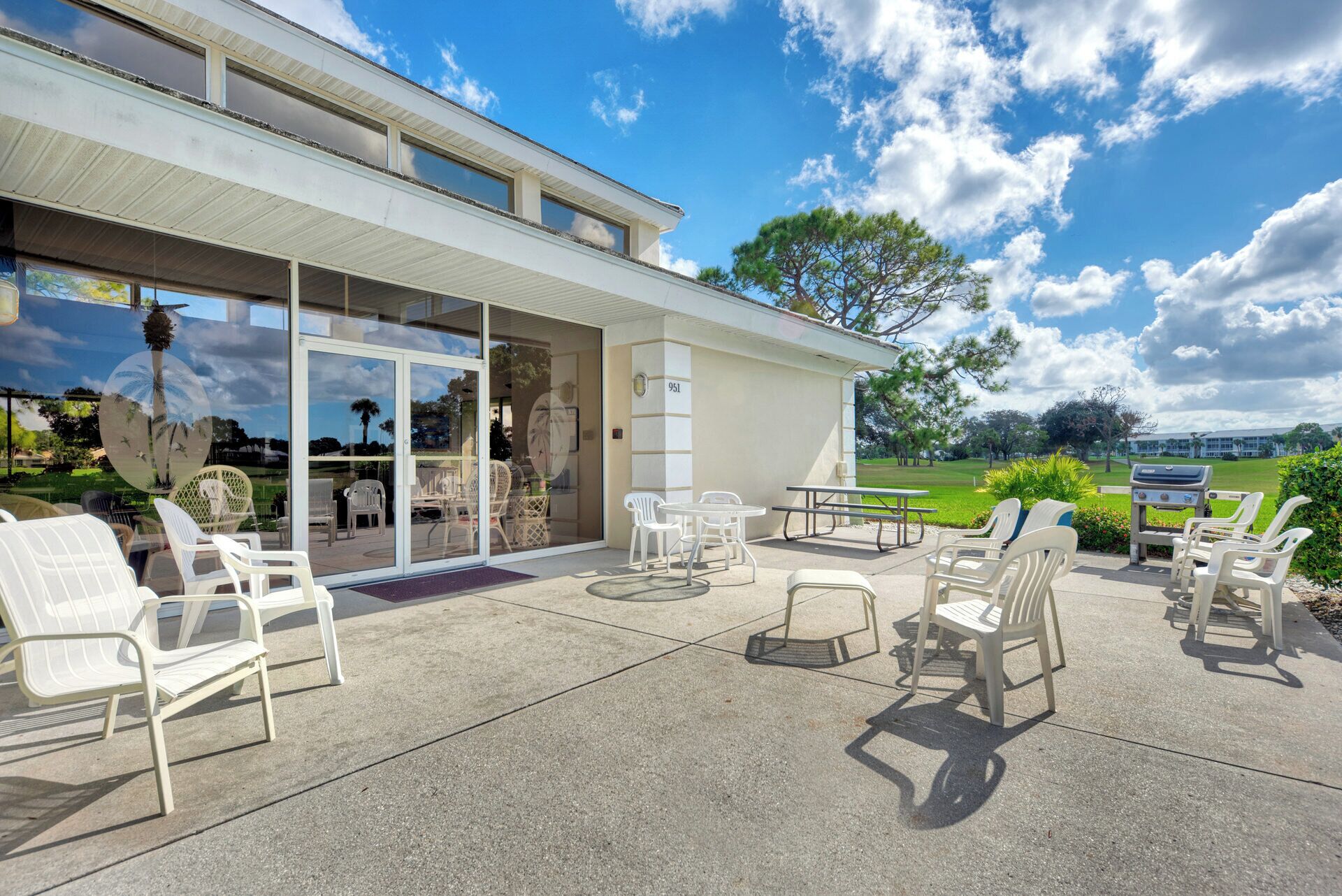 Um pátio coberto ao ar livre com cadeiras e mesas de plástico branco, uma porta de vidro que dá para uma área de piscina e vista para um campo de golfe.