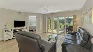 Un salon spacieux avec un grand canapé en cuir, un téléviseur à écran plat et une table basse en verre. Une porte-fenêtre coulissante donne accès à un patio aménagé avec du mobilier d’extérieur.