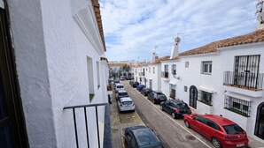Uma rua estreita ladeada por edifícios brancos, carros estacionados e um céu claro.