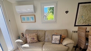 Een woonkamer met een bank, een ronde houten salontafel, een tapijt met dessin, een ingelijste kaart en een raam met uitzicht op groen.