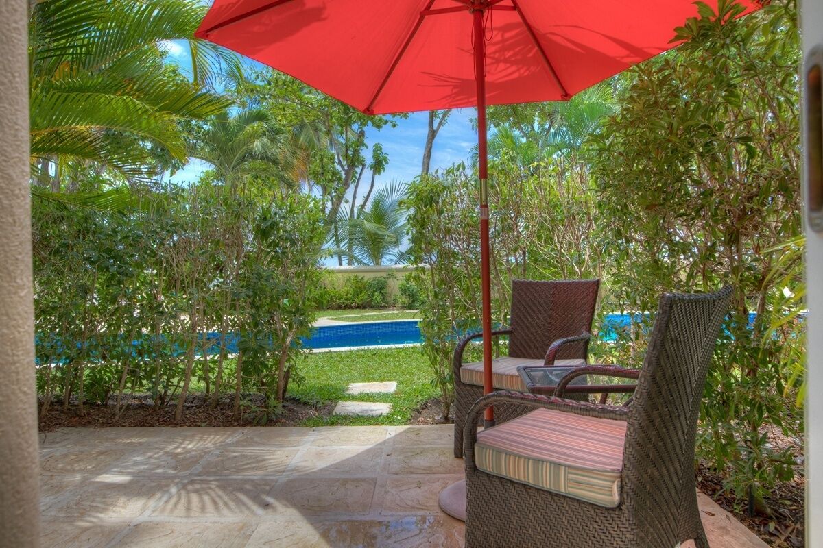 Un patio con mobiliario de mimbre y un paraguas rojo, con vistas a una piscina y abundante vegetación.