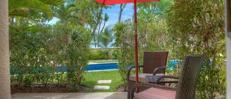 Un patio con mobiliario de mimbre y un paraguas rojo, con vistas a una piscina y abundante vegetación.