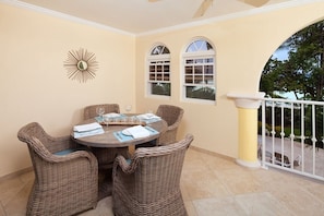 A covered patio with a round table and wicker chairs, two windows, and a decorative sunburst wall piece. - Sapphire Beach 116 2 Bedroom Apts (St. Lawrence Gap)