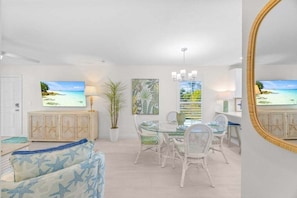 A spacious living room with a glass dining table, white wicker chairs, a large mirror, and a view of the ocean through a window. - Seashells Unit 23 2 Bedroom Condo (Sanibel)