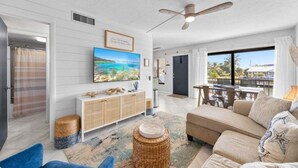 Ein modernes Wohnzimmer mit einem beige farbenen Sofa, einem an der Wand befestigten Fernseher und einem großen Fenster mit Blick ins Freie.