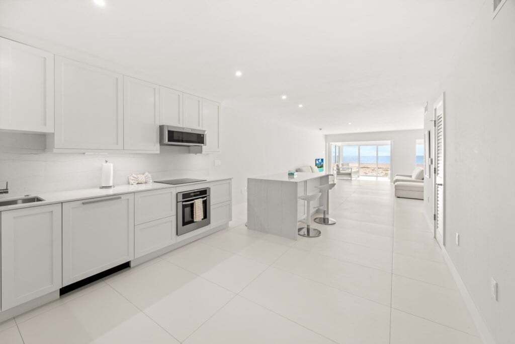 A modern kitchen with white cabinetry, a central island with bar stools, and a view of the beach through large windows.